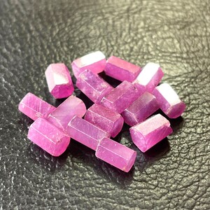 May include: A collection of raw, hexagonal-shaped ruby gemstones. The gemstones are a vibrant pinkish-red color with white tips, and are scattered on a dark, textured surface. The gemstones are uncut and appear to be natural.