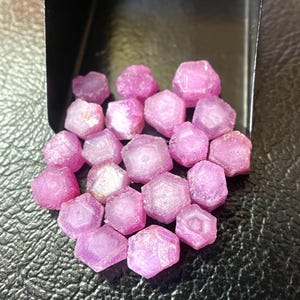 May include: A collection of small, faceted, pink gemstones. The gems have a hexagonal shape and a translucent appearance. They are displayed in a black tray against a dark background.