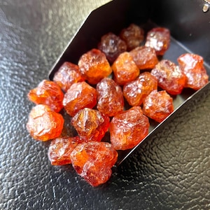 May include: A collection of raw, orange-red gemstones, possibly garnets, displayed in a small, dark metal container. The gemstones have a rough, crystalline texture and varying shapes. The background is a textured, dark surface.