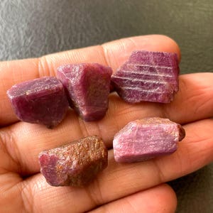 Peut inclure: Cinq pierres précieuses de rubis brutes de formes et tailles variées. Les rubis présentent une teinte rouge foncé, certains affichant une teinte brunâtre. Les pierres sont tenues dans une main, mettant en valeur leur aspect naturel et non poli.