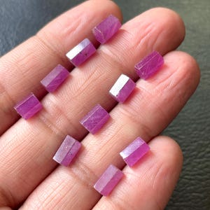 May include: A collection of small, faceted ruby gemstones. The gemstones are a vibrant pinkish-red color and rectangular in shape. The gems are held in a hand, showcasing their polished surfaces and natural variations.