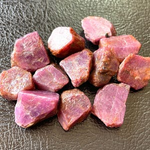 May include: A cluster of raw ruby gemstones in various sizes and forms. The rubies exhibit a spectrum of pink and red tones, with a textured, natural look. The gemstones are arranged on a dark, textured surface.
