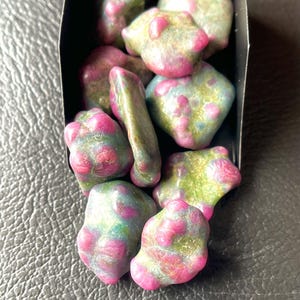 Könnte beinhalten: Eine Sammlung von Trommelsteinen in einer schwarzen Box. Die Steine weisen eine Mischung aus Farben auf, mit grünen und blauen Basen und leuchtend rosa Flecken. Die Steine haben eine unregelmäßige Form.