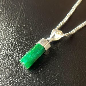 Puede incluir: Un collar con un colgante rectangular verde brillante y una cadena plateada. El colgante es de un color verde esmeralda vibrante, con una tapa plateada. La cadena es una delicada cadena de eslabones plateada.
