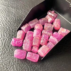 May include: A collection of raw, uncut ruby gemstones. The gemstones are a vibrant pinkish-red color and have a rough, textured surface. They are contained within a black tray, showcasing their natural beauty and potential for jewelry making.