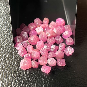 Könnte beinhalten: Eine Sammlung roher, ungeschliffener Rubin-Edelsteine. Die Rubine haben eine lebendige, rötlich-rosa Farbe und eine raue, natürliche Textur. Sie werden in einer kleinen, schwarzen Schale vor dunklem Hintergrund präsentiert. Geeignet für die Schmuckherstellung.