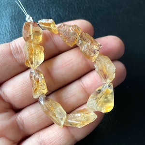 Könnte beinhalten: Ein Armband aus grob geschnittenen Citrin-Edelsteinen. Die Steine haben eine durchscheinende gelb-orange Farbe mit unregelmäßigen Formen und sichtbaren Einschlüssen. Das Armband ist an einem dünnen, weißen Band aufgereiht.
