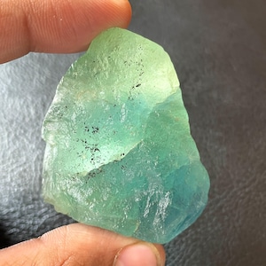 Puede incluir: Una gema de fluorita natural en bruto con un degradado de colores. La piedra pasa de un verde claro en la parte superior a un verde azulado oscuro en la parte inferior. La superficie tiene un aspecto rugoso y texturizado con pequeñas inclusiones negras.
