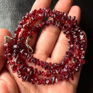 May include: A red garnet chip necklace with a silver clasp. The necklace is made of small, irregularly shaped, deep red garnet stones. The necklace is displayed on a hand.