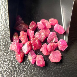 Könnte beinhalten: Eine Sammlung roher, ungeschliffener Rubin-Edelsteine. Die Steine variieren in Größe und Form und weisen einen leuchtenden rosa bis tiefroten Farbton auf. Die Edelsteine werden in einem kleinen, dunklen Behälter vor dunklem Hintergrund präsentiert.