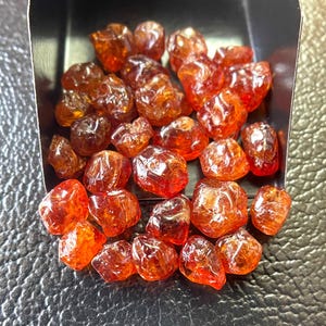 May include: A collection of raw, uncut, orange-red gemstones. The gems are irregularly shaped with a translucent quality, displayed in a small, dark tray. The stones have a polished appearance, reflecting light and showcasing their natural color variations.