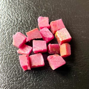 Peut inclure: Un ensemble de pierres précieuses rubis roses brutes. Les pierres sont taillées grossièrement, avec des formes variées, dont des cubes et des éclats. Les morceaux de rubis naturels sont dispersés sur une surface sombre et texturée, mettant en valeur leur couleur et leur texture.