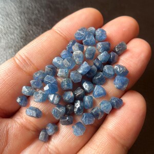 May include: A handful of small, faceted blue gemstones. The stones vary in shade from light to dark blue, with a slightly rough, natural texture. The gemstones are held in an open hand, with a dark background.