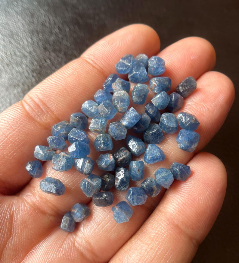 May include: A handful of small, faceted blue gemstones. The stones vary in shade from light to dark blue, with a slightly rough, natural texture. The gemstones are held in an open hand, with a dark background.