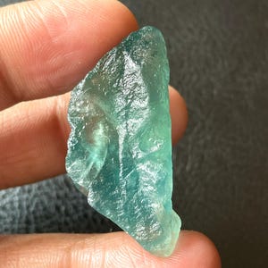 Puede incluir: Una gema translúcida de color verde azulado, de forma irregular. La piedra tiene una superficie rugosa y texturizada y diferentes tonos de azul y verde. La gema se sostiene entre los dedos, mostrando su forma natural y variaciones de color.