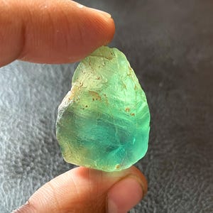 Puede incluir: Una gema de fluorita en bruto, translúcida, con gradientes de tonos verdes y azules. La piedra se sostiene entre los dedos, mostrando su forma irregular y textura natural. La gema tiene una apariencia cruda y orgánica.