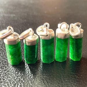 Puede incluir: Cinco colgantes de piedras preciosas verdes con tapas y bucles plateados. Los colgantes cilíndricos tienen lados facetados, mostrando un tono verde vibrante. Las tapas plateadas tienen un acabado pulido.