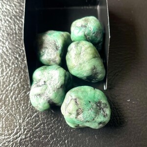 May include: A collection of rough emerald gemstones in a small black box. The emeralds are a vibrant green color with dark inclusions, showcasing their natural, unpolished state. The gemstones are displayed against a dark background.