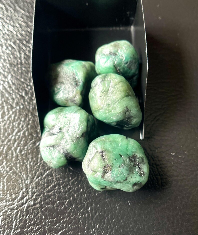 May include: A collection of rough emerald gemstones in a small black box. The emeralds are a vibrant green color with dark inclusions, showcasing their natural, unpolished state. The gemstones are displayed against a dark background.