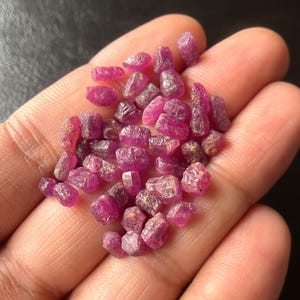 Könnte beinhalten: Nahaufnahme einer Handvoll roher, ungeschliffener Rubin-Edelsteine. Die Rubine haben eine tiefrosa-rote Farbe mit einem leicht durchscheinenden Aussehen. Die Edelsteine variieren in Größe und Form und haben eine natürliche, unpolierte Textur.