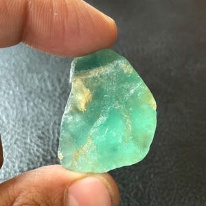 Puede incluir: Una gema translúcida de color verde azulado, de forma irregular, con áreas de color marrón claro y amarillo. La piedra se sostiene entre los dedos, mostrando su forma natural y en bruto. La gema tiene un aspecto liso y pulido.