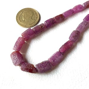 May include: A strand of rough-cut, cylindrical ruby beads in varying shades of pink and red. The beads are displayed next to a gold coin for size comparison. The gemstones have a natural, unpolished appearance.