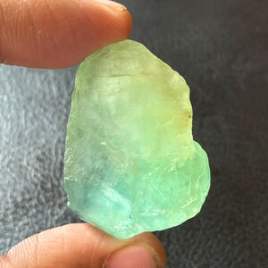 May include: A raw, translucent gemstone displaying a colour gradient, shifting from pale yellow to vivid green and touches of blue. The stone is held between fingers, highlighting its natural, irregular form and texture.