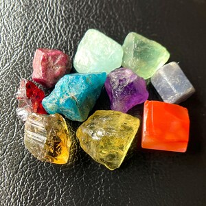 May include: A collection of colorful, raw gemstones in various shapes and sizes. The stones include shades of red, blue, green, purple, yellow, and orange. The gemstones are displayed on a dark, textured surface.