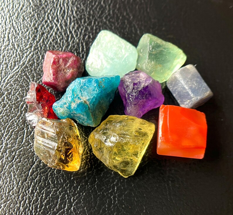 May include: A collection of colorful, raw gemstones in various shapes and sizes. The stones include shades of red, blue, green, purple, yellow, and orange. The gemstones are displayed on a dark, textured surface.