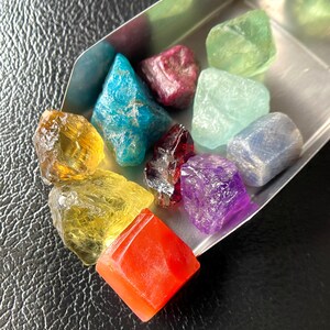 May include: A collection of colorful, raw gemstones in a small metal tray. The stones vary in color, including yellow, blue, red, purple, and green. The gemstones have a rough, natural texture and are displayed against a dark background.