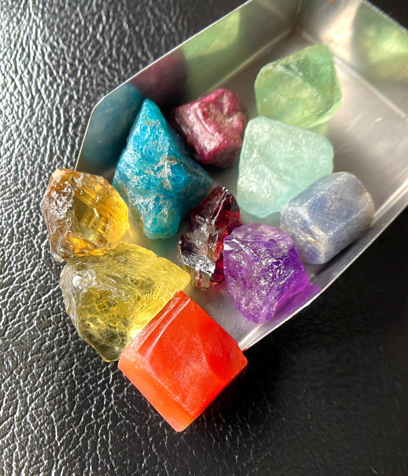 May include: A collection of colorful, raw gemstones in a small metal tray. The stones vary in color, including yellow, blue, red, purple, and green. The gemstones have a rough, natural texture and are displayed against a dark background.