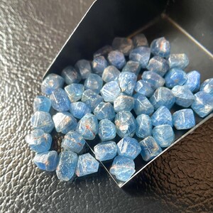 May include: A collection of small, light blue gemstones with irregular shapes. The stones have a translucent quality, with some showing hints of red or orange. They are contained within a black tray, suggesting they are for sale or display.