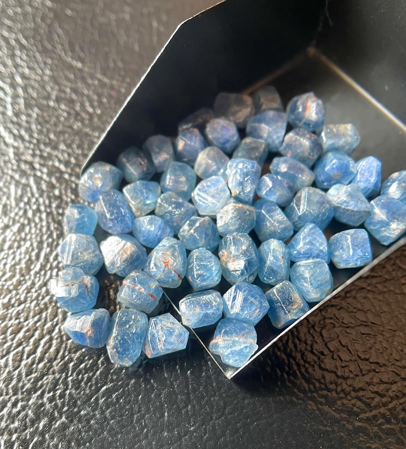 May include: A collection of small, light blue gemstones with irregular shapes. The stones have a translucent quality, with some showing hints of red or orange. They are contained within a black tray, suggesting they are for sale or display.
