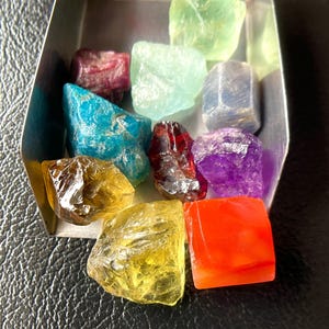 May include: A collection of colorful, raw gemstones in a small, silver tray. The stones vary in color, including green, blue, red, yellow, and purple. The gemstones are displayed on a black surface.
