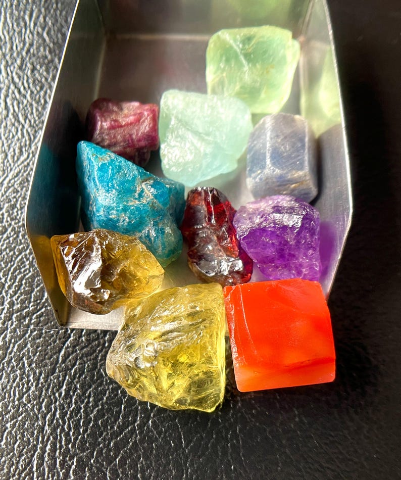 May include: A collection of colorful, raw gemstones in a small, silver tray. The stones vary in color, including green, blue, red, yellow, and purple. The gemstones are displayed on a black surface.