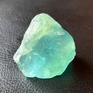 Puede incluir: Un cristal de fluorita en bruto y natural, con una mezcla de tonos azules y verdes. La gema tiene una forma rugosa e irregular y un aspecto translúcido, mostrando su textura natural y variaciones de color. Un ejemplo vibrante de belleza mineral natural.