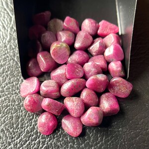 Può includere: Una collezione di pietre preziose di rubino burattate in un piccolo contenitore nero. Le pietre preziose sono di un colore rosa-rossastro intenso, con una consistenza liscia e lucida. Le pietre preziose sono di varie dimensioni e forme.