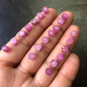 Pode incluir: Uma coleção de pequenas pedras preciosas de rubi em bruto. As pedras preciosas são de uma cor rosa-avermelhada vibrante e têm uma aparência natural e não polida. Estão espalhadas por uma mão, mostrando suas formas e texturas individuais. O fundo é escuro e neutro.