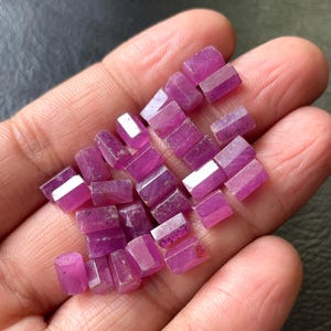 May include: A collection of small, faceted, pink-purple gemstones. The gemstones are rectangular with angled edges, displaying a range of shades from light to deep magenta. The gems are held in a hand, with a dark background.