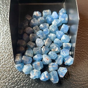 May include: A collection of raw, uncut blue gemstones. The stones are a mix of light and dark blue hues, with some showing hints of red. They are contained within a small, dark tray, resting on a textured black surface. The gemstones have a rough, natural appearance.