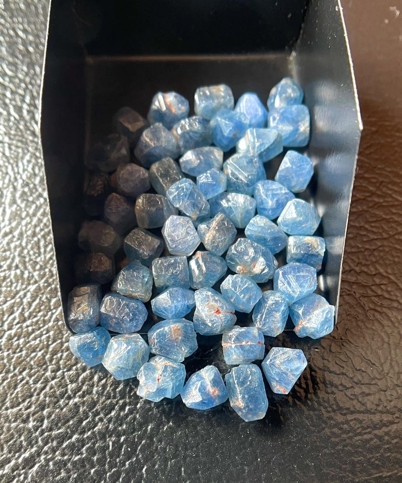 May include: A collection of raw, uncut blue gemstones. The stones are a mix of light and dark blue hues, with some showing hints of red. They are contained within a small, dark tray, resting on a textured black surface. The gemstones have a rough, natural appearance.