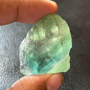 Puede incluir: Un cristal de fluorita en bruto, con una mezcla de tonos verdes y azules. La piedra natural se sostiene entre los dedos, mostrando su cualidad translúcida y forma irregular. La superficie del cristal presenta texturas y variaciones de color.