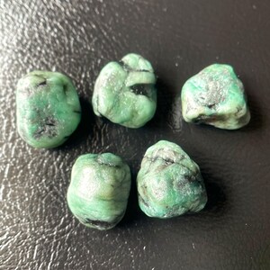 May include: Five raw emerald gemstones with a vibrant green hue and dark inclusions. The stones vary in size and shape, showcasing natural textures. The gemstones are displayed on a dark surface.