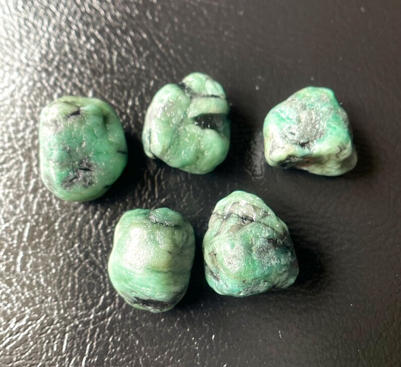 May include: Five raw emerald gemstones with a vibrant green hue and dark inclusions. The stones vary in size and shape, showcasing natural textures. The gemstones are displayed on a dark surface.