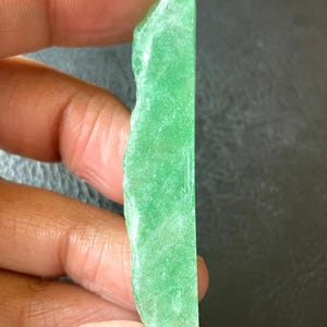 Puede incluir: Una vibrante rebanada de piedra preciosa verde crudo. La piedra tiene un borde áspero e irregular en un lado y un borde liso y pulido en el otro. El color es un verde brillante y translúcido, con una superficie ligeramente texturizada. La piedra preciosa se sostiene entre los dedos.