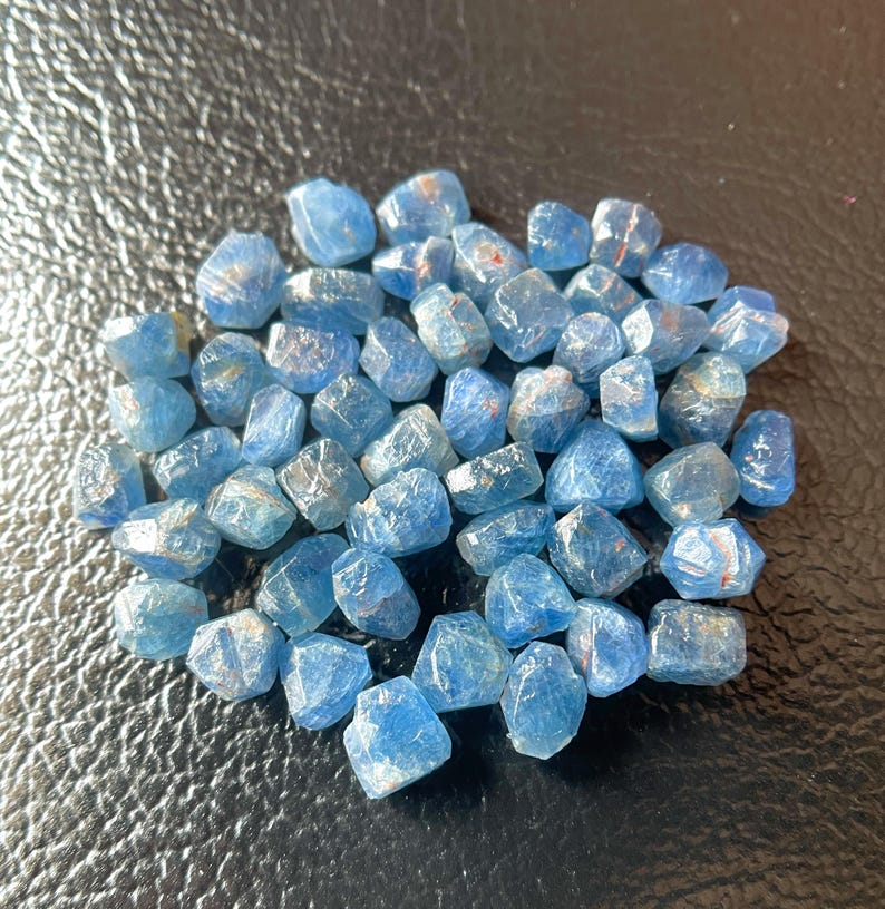 May include: A collection of small, faceted blue gemstones. The stones have a translucent quality with some showing internal inclusions. The gems are scattered on a dark, textured surface, creating a contrast that highlights their color and form.