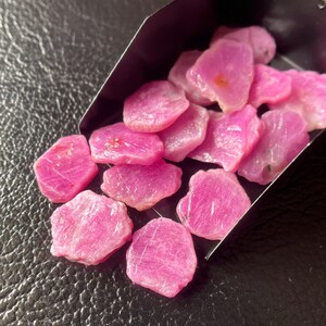 Op de afbeelding: Een verzameling ruwe, roze robijn edelstenen. De ongeslepen stenen hebben een natuurlijke, onregelmatige vorm en een levendige, verzadigde kleur. De edelstenen worden getoond in een donkere metalen schep tegen een donkere achtergrond.