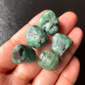 May include: A collection of six raw emerald stones. The emeralds are a deep green color with some black inclusions. The stones are irregularly shaped and appear to be natural, unpolished specimens. The image is a close-up, showing the texture and color variations.