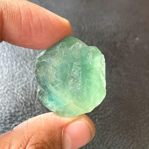 Puede incluir: Una gema de fluorita en bruto, translúcida, en tonos verdes y azules. La piedra se sostiene entre los dedos, mostrando su forma natural irregular y su estructura cristalina. La gema tiene una textura ligeramente rugosa.