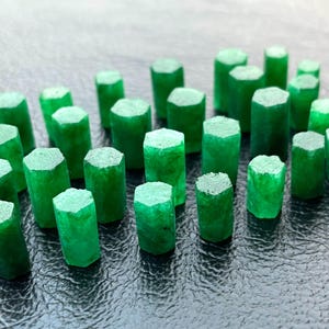 May include: A collection of vibrant green emerald crystals, each with a hexagonal top and a cylindrical body. The gemstones are arranged closely together, showcasing their rich color and natural formations.